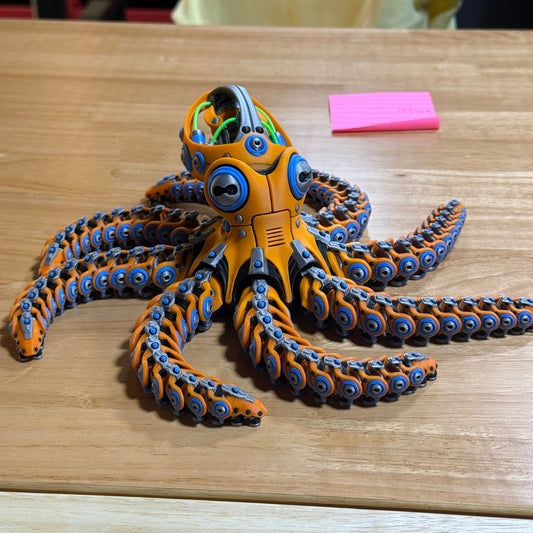 A 3D-printed articulated octopus toy with orange, blue, and black segments, mounted on a light wood panel next to a pink sticky note.