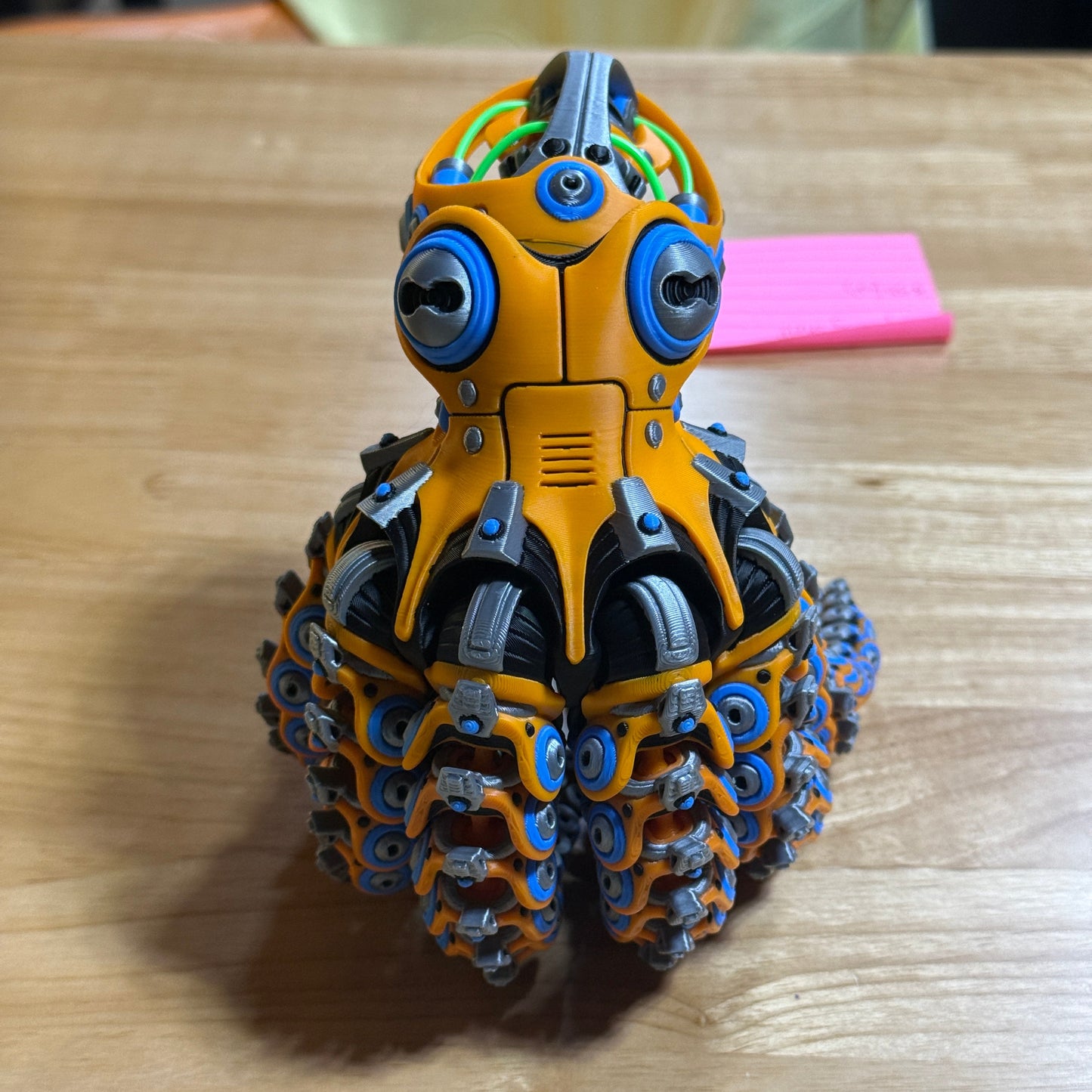 A small orange and blue robotic toy or figurine with multiple circular eyes and mechanical details, placed on a wooden tabletop with a pink sticky note in the background.